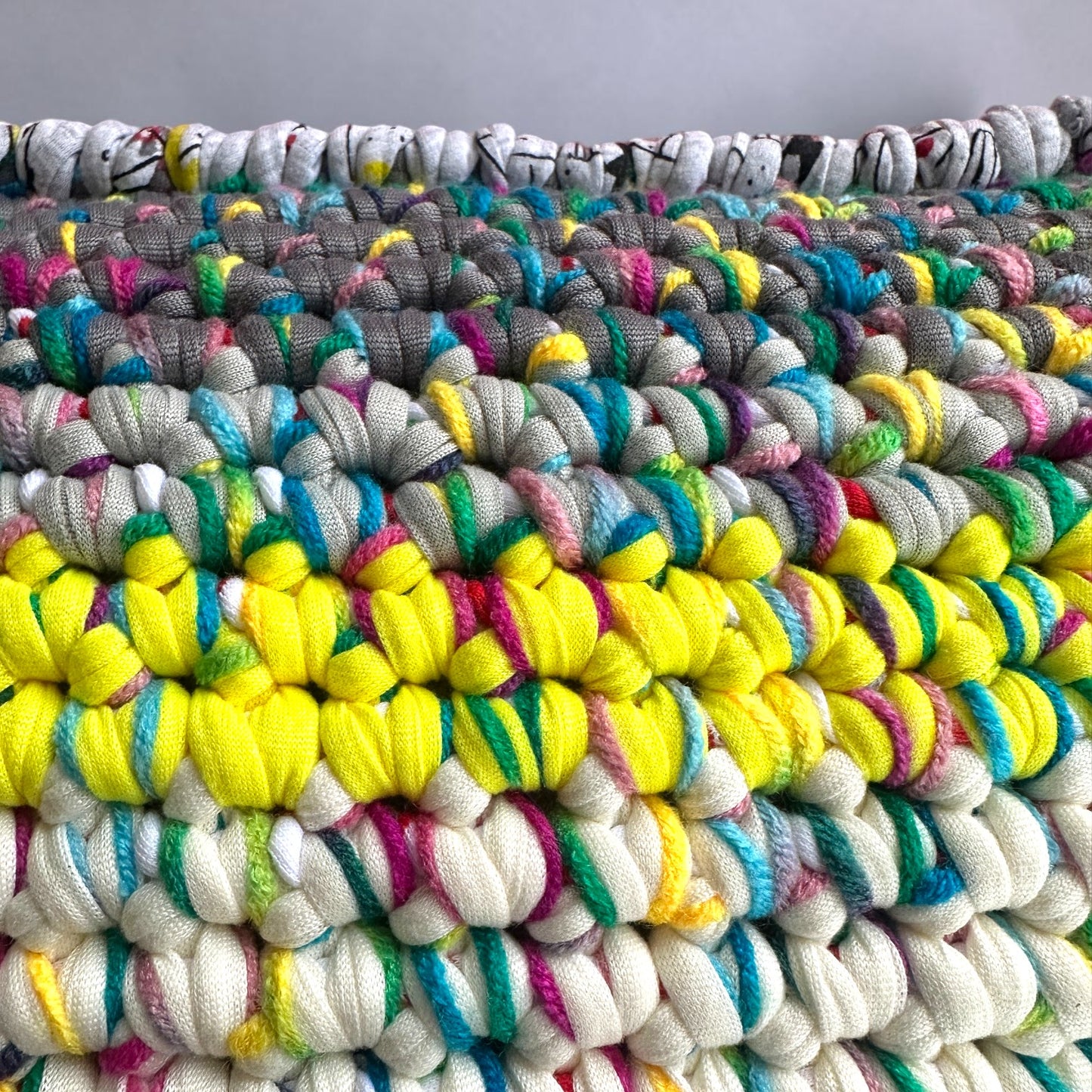 Nahaufnahme der Coiled-Crochet-Struktur des Kissens Sundown Grey, Detail der oberen Kante mit Strichzeichnungen auf hellgrauem Grund und HOOKamSEE-Label