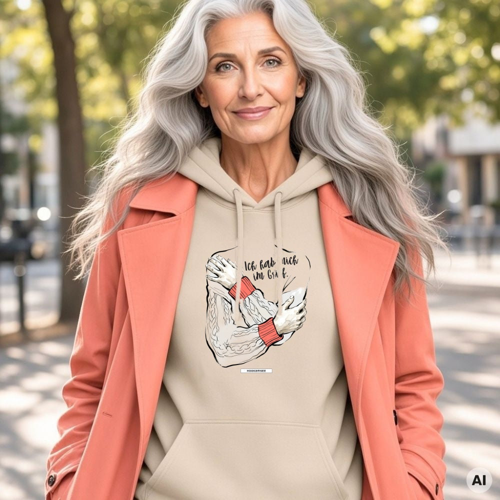 Frau mit langen grauen Haaren und korallfarbenem Mantel trägt den HOOKamSEE Hoodie Ich hab mich im Griff. in Sand unter Bäumen in der Stadt