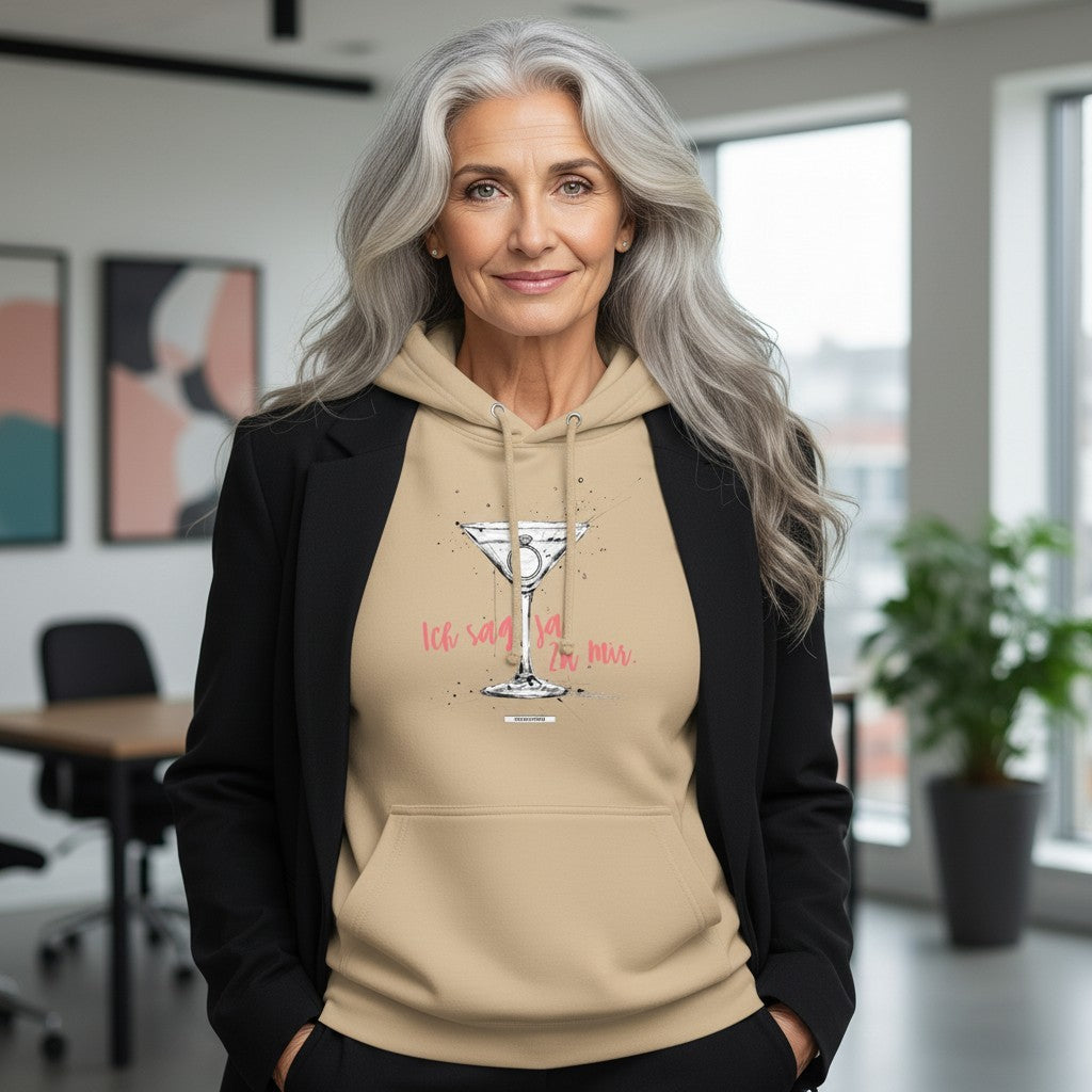 Frau mit grauen Haaren trägt sandfarbenen Hoodie mit Cocktailglas-Motiv und Schriftzug Ich sag ja. Zu mir. in Koralle, dazu schwarzer Blazer, modernes Büro im Hintergrund