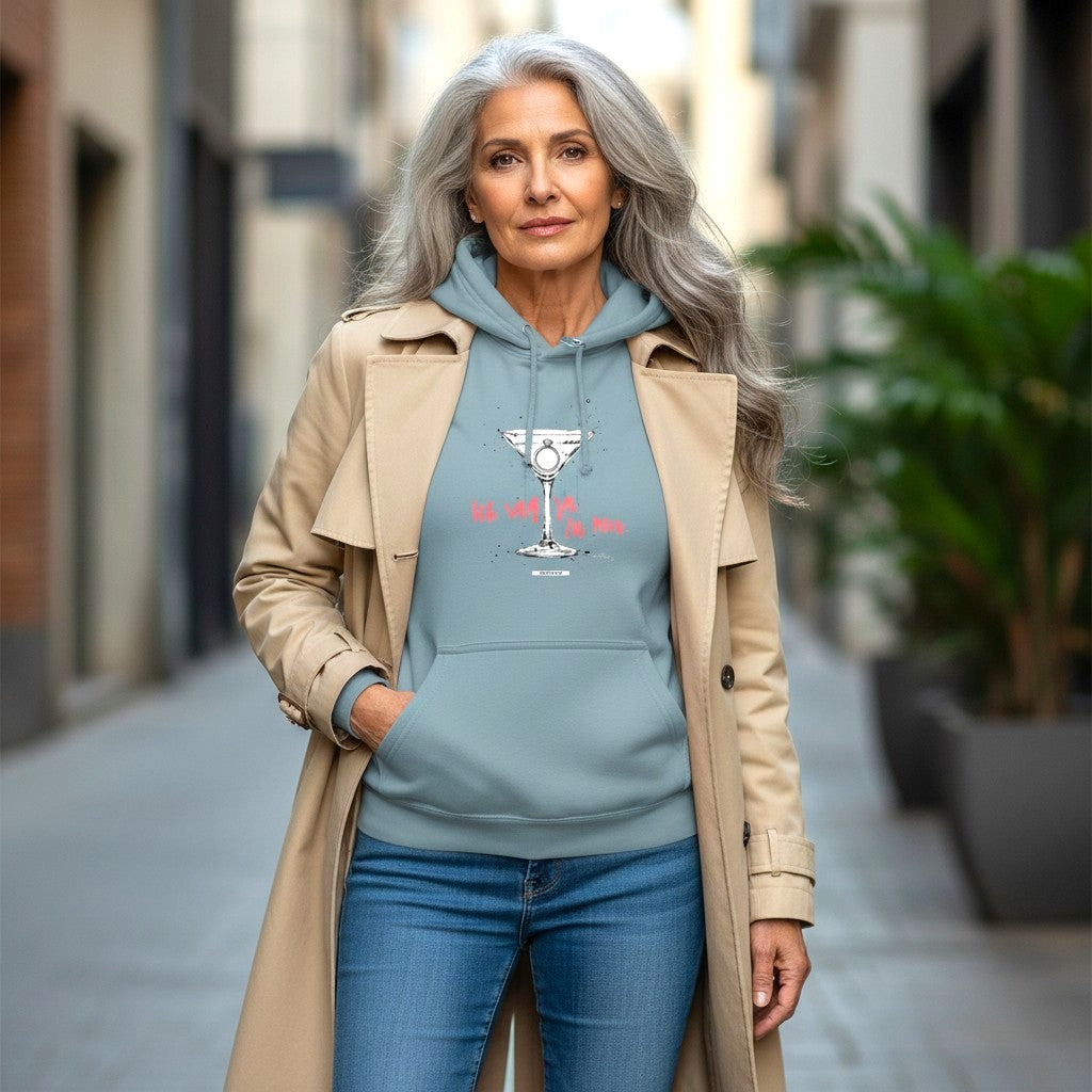 Frau mit grauen Haaren trägt nebelblauen Hoodie mit Cocktailglas-Motiv und Schriftzug Ich sag ja. Zu mir. in Koralle, dazu beiger Trenchcoat und Jeans, städtische Straßenszene
