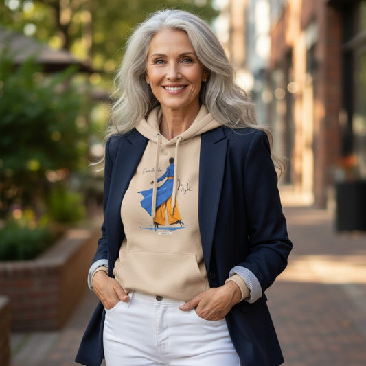 Frau mit langen, grauen Haaren trägt HOOKamSEE Hoodie Karma klärt das. Ich nicht. in Sand, kombiniert mit dunkelblauem Blazer und weißer Jeans, auf einer Straße mit grünen Bäumen im Hintergrund