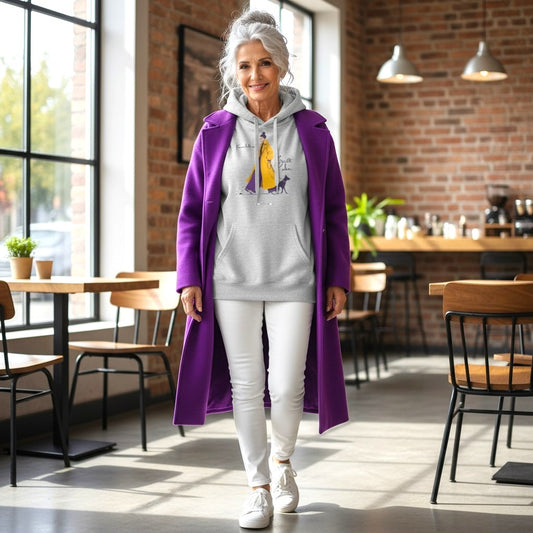 Frau mittleren Alters in grauem HOOKamSEE Hoodie „Karma bellt nicht. Beißt aber." mit Hunde-Print, kombiniert mit lila Mantel und weißer Jeans, in einem Café mit Backsteinwand.