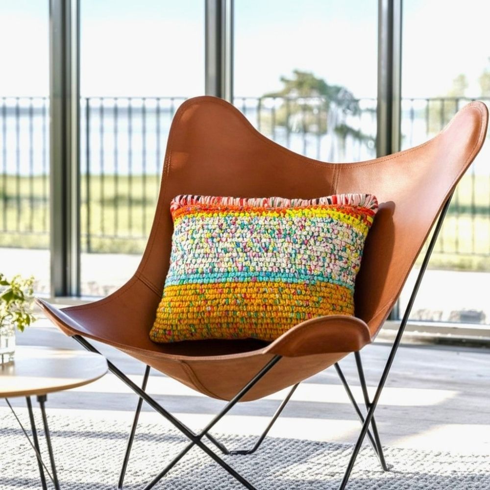 Kissen Golden Carnival auf einem braunen Leder-Butterfly-Chair vor einer Fensterfront mit Seeblick