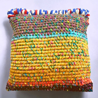 Coiled-Crochet-Kissen Saffron Clay 45×45 cm mit Farbverlauf von Taupe-Braun über Türkis zu Ocker-Gelb, unterer Rand in Rostorange-Braun, oberer Rand mit rot-weiß-blauer Bändchengarn-Kante, freigestellt auf hellgrauem Hintergrund