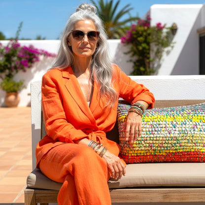 Frau in orangefarbenem Leinenanzug lehnt neben Kissen Sundown Grey auf Outdoor-Bank vor mediterraner Kulisse