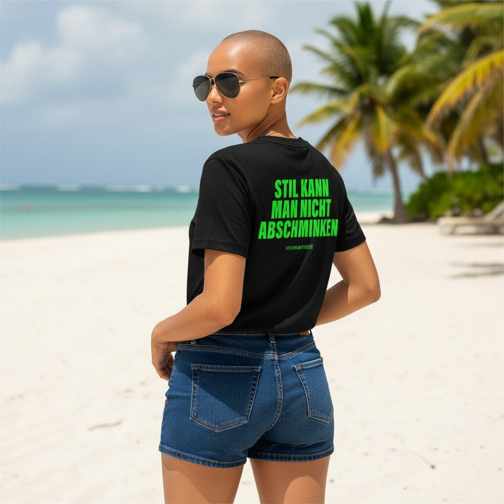 Frau mit Glatze und Sonnenbrille am Karibikstrand trägt schwarzes Unisex-T-Shirt mit neongrünem Aufdruck Stil kann man nicht abschminken auf dem Rücken, HOOKamSEE, Jeans-Shorts.