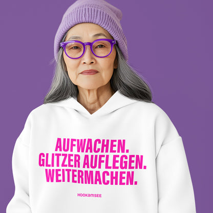 Frau trägt weißen Unisex Hoodie mit pinkem Statement „AUFWACHEN. GLITZER AUFLEGEN. WEITERMACHEN.“ von HOOKamSEE